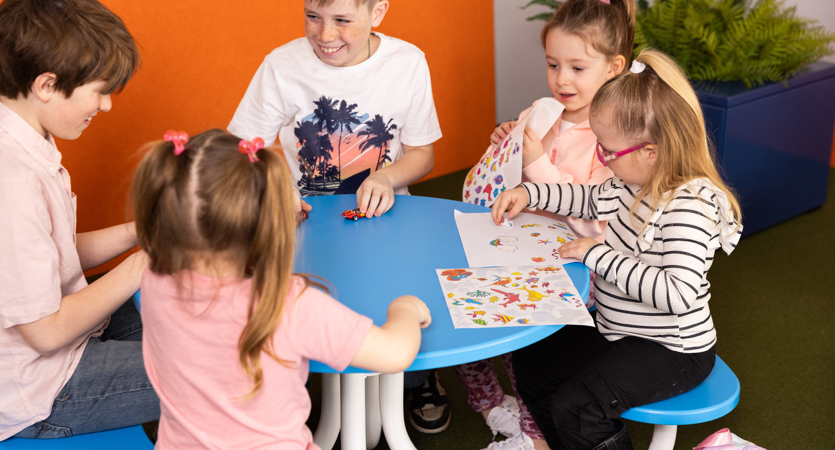Quatre enfants sont assis autour d'une table bleue, s'amusant avec des autocollants et du papier, tandis qu'un autre enfant se tient à proximité. Il semble s'agir d'une salle de classe ou d'une aire de jeux.