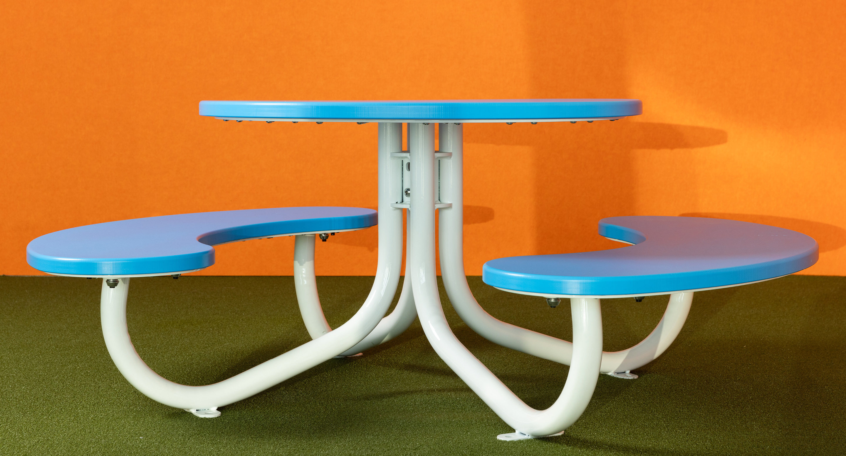 Table de pique-nique ronde bleue avec deux bancs courbés sur des pieds en métal blanc, sur un mur orange et un sol vert.