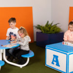 Quatre enfants jouent à un jeu de société sur une table ronde bleue, tandis que trois autres enfants interagissent avec un jeu sur un cube bleu et blanc dans une salle de jeux colorée.
