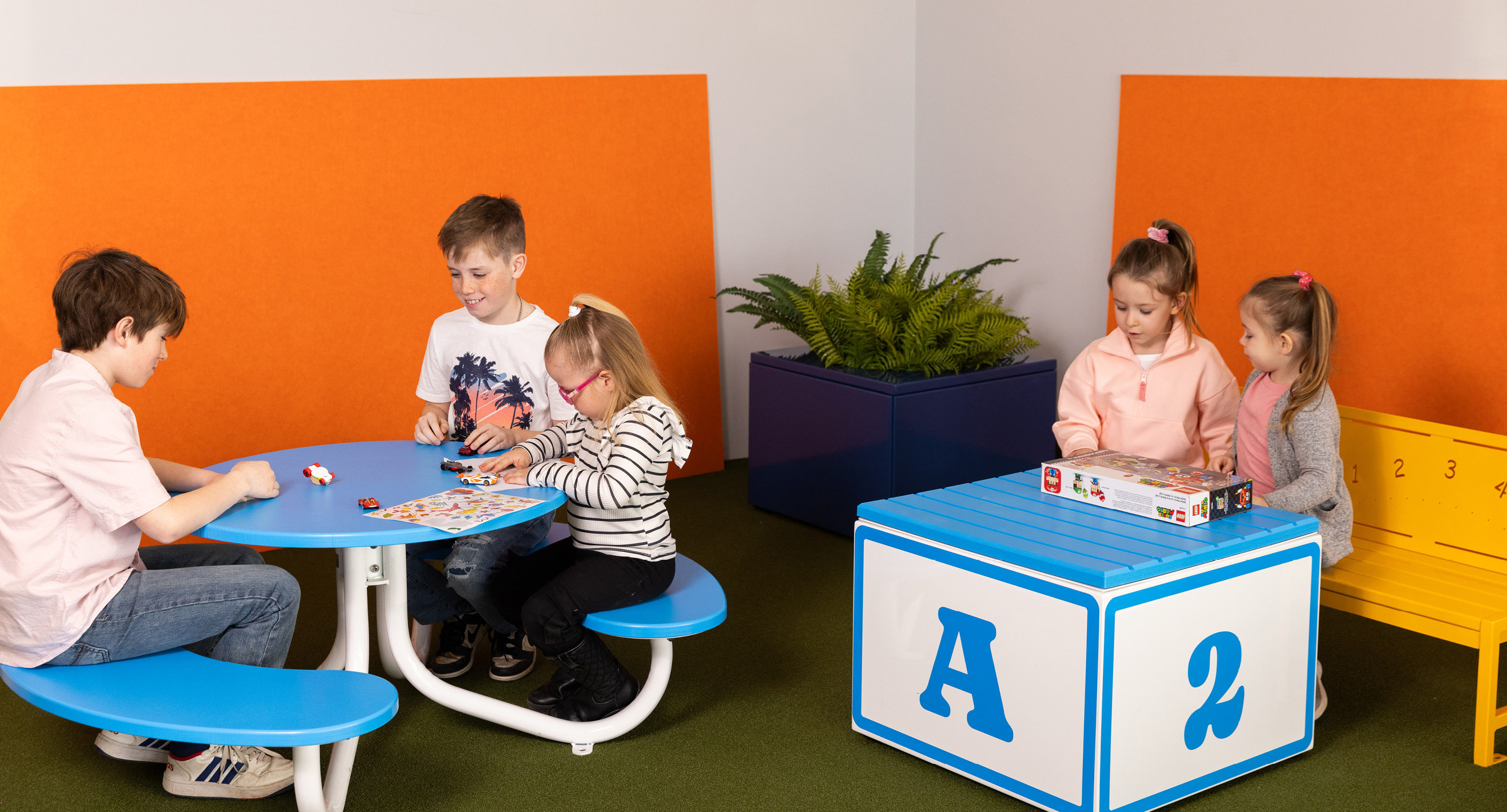 maglin_childrens-fava-picnic-tables-5 Quatre enfants jouent à un jeu de société sur une table ronde bleue, tandis que trois autres enfants interagissent avec un jeu sur un cube bleu et blanc dans une salle de jeux colorée.