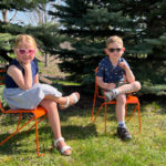 Deux jeunes enfants sont assis sur des chaises orange en plein air et portent des lunettes de soleil. Ils sont placés sur de l'herbe avec des pins en arrière-plan par une journée ensoleillée.