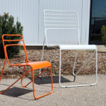 Une chaise métallique orange de la taille d'un enfant et une chaise métallique blanche plus grande sont placées à l'extérieur, sur le trottoir, près de petits arbustes et d'un mur d'enceinte.