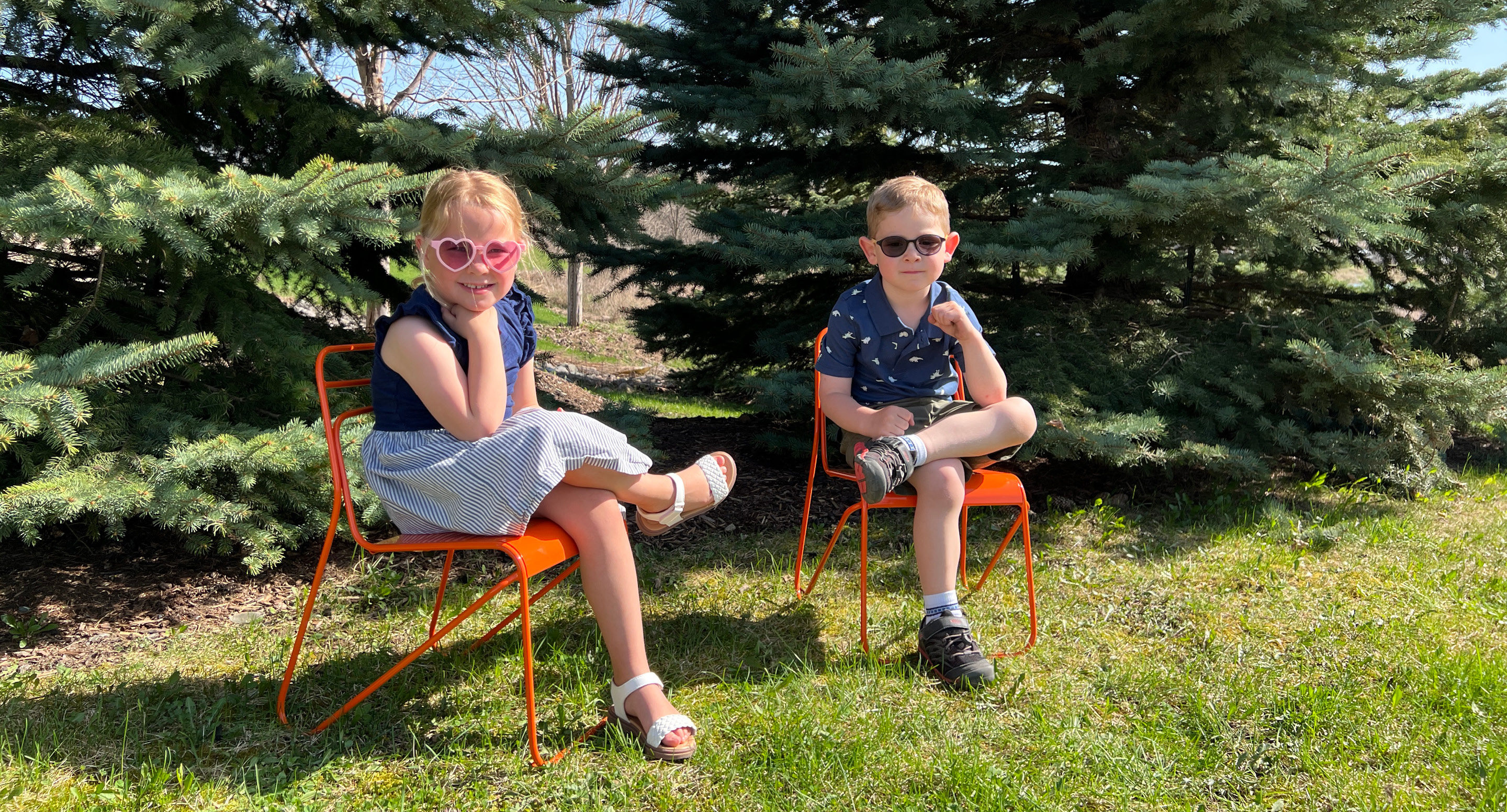 maglin_childrens-kontur-chair-1 Deux jeunes enfants sont assis sur des chaises orange en plein air et portent des lunettes de soleil. Ils sont placés sur de l'herbe avec des pins en arrière-plan par une journée ensoleillée.
