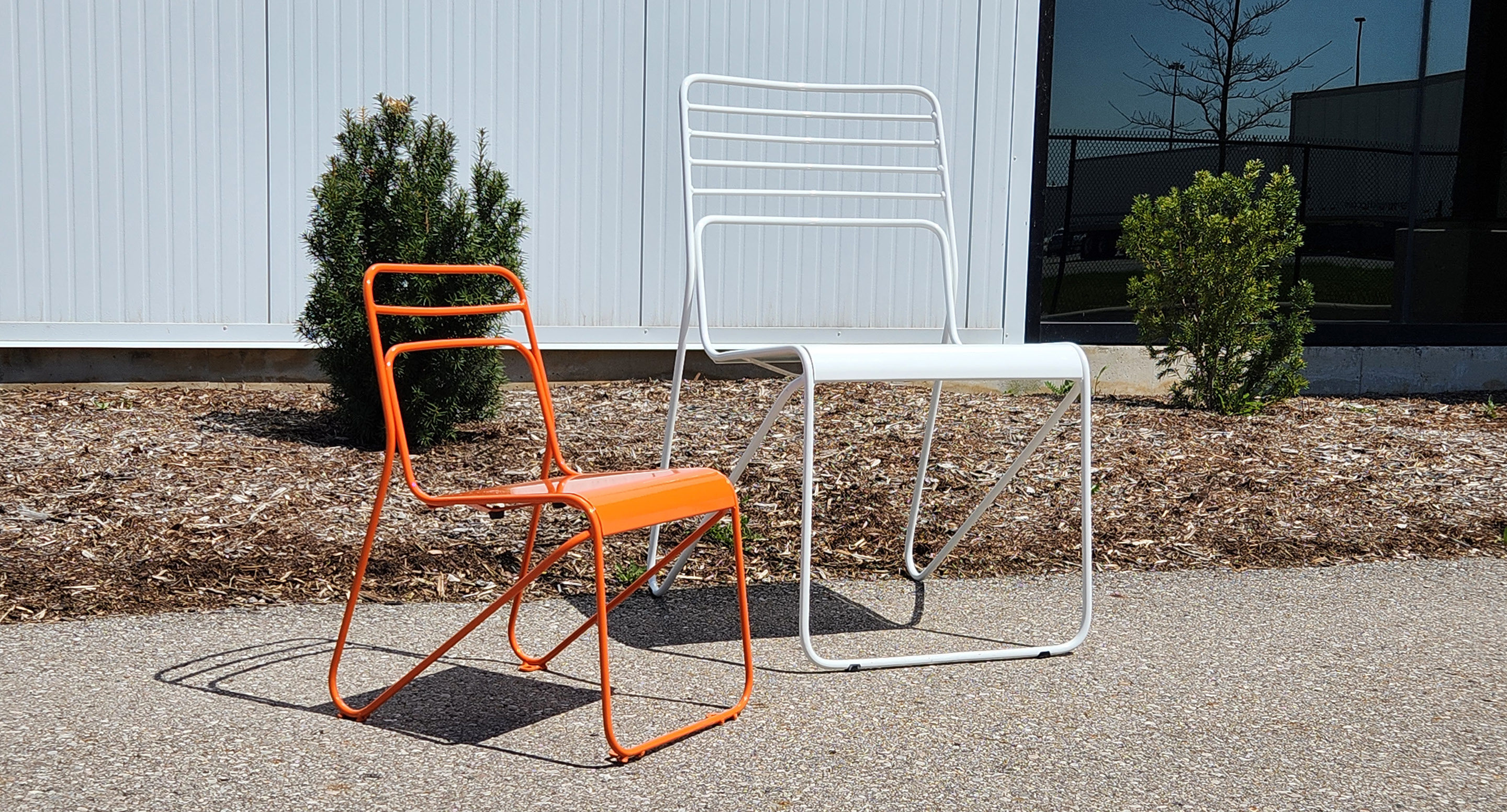 maglin_childrens-kontur-chair-1 Une chaise métallique orange de la taille d'un enfant et une chaise métallique blanche plus grande sont placées à l'extérieur, sur le trottoir, près de petits arbustes et d'un mur d'enceinte.