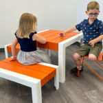 Deux jeunes enfants sont assis à une table orange et blanche ; une fille est assise sur un banc tandis que le garçon est assis sur une chaise orange, tous deux se faisant face dans une pièce lumineuse au sol gris.