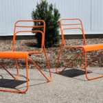 Deux chaises modernes en métal orange sont placées sur une surface pavée à l'extérieur avec du paillis, un petit buisson et une clôture blanche en arrière-plan.