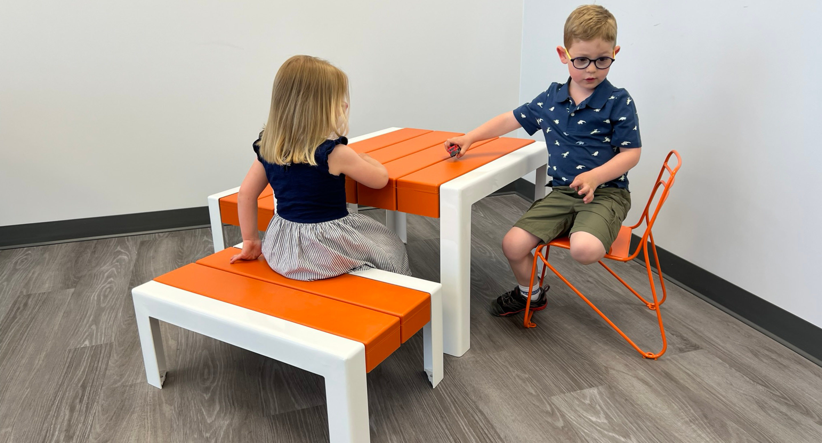 maglin_childrens-kontur-chair-2 Deux jeunes enfants sont assis à une table orange et blanche ; une fille est assise sur un banc tandis que le garçon est assis sur une chaise orange, tous deux se faisant face dans une pièce lumineuse au sol gris.