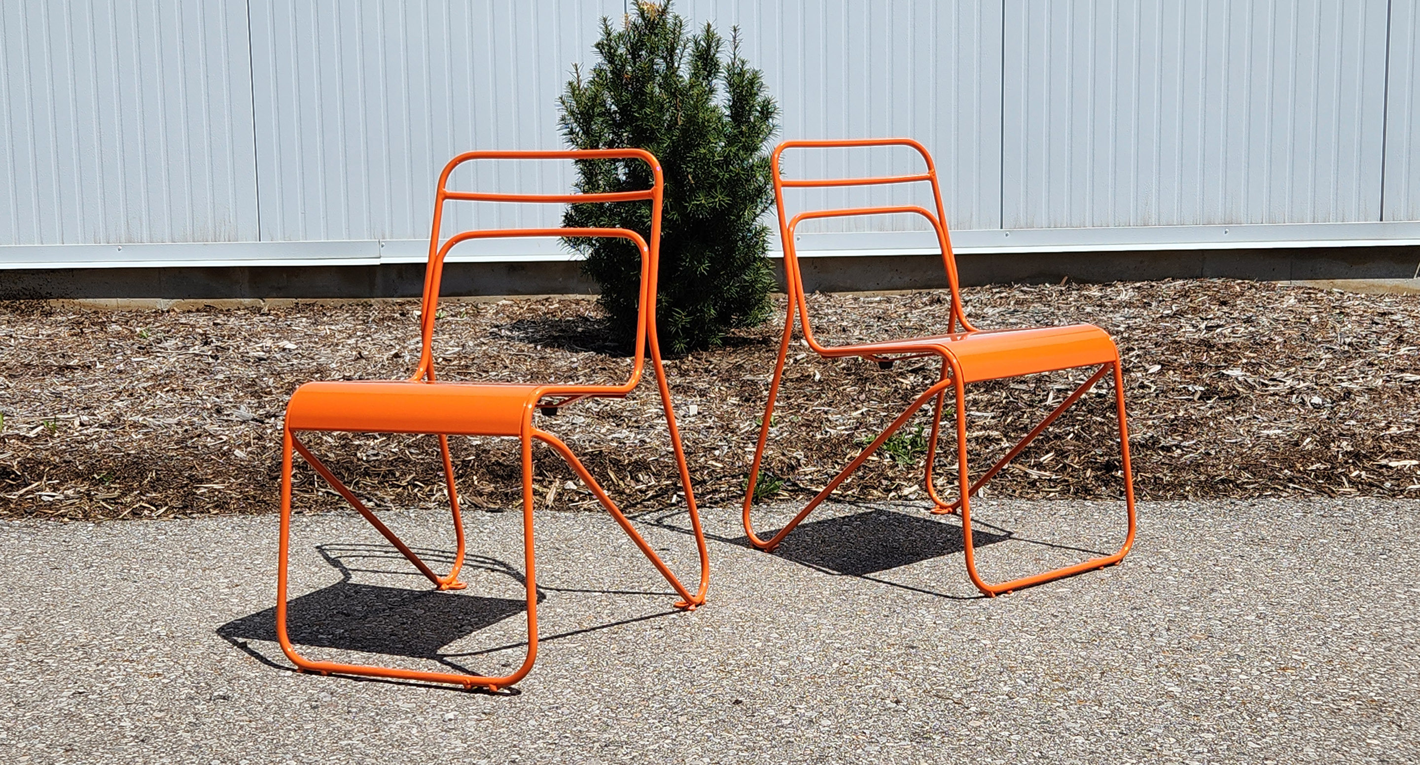 maglin_childrens-kontur-chair-2 Deux chaises modernes en métal orange sont placées sur une surface pavée à l'extérieur avec du paillis, un petit buisson et une clôture blanche en arrière-plan.