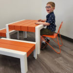 Un jeune garçon sourit alors qu'il est assis à une table orange et blanche posée sur un sol en bois dans une pièce aux murs blancs.