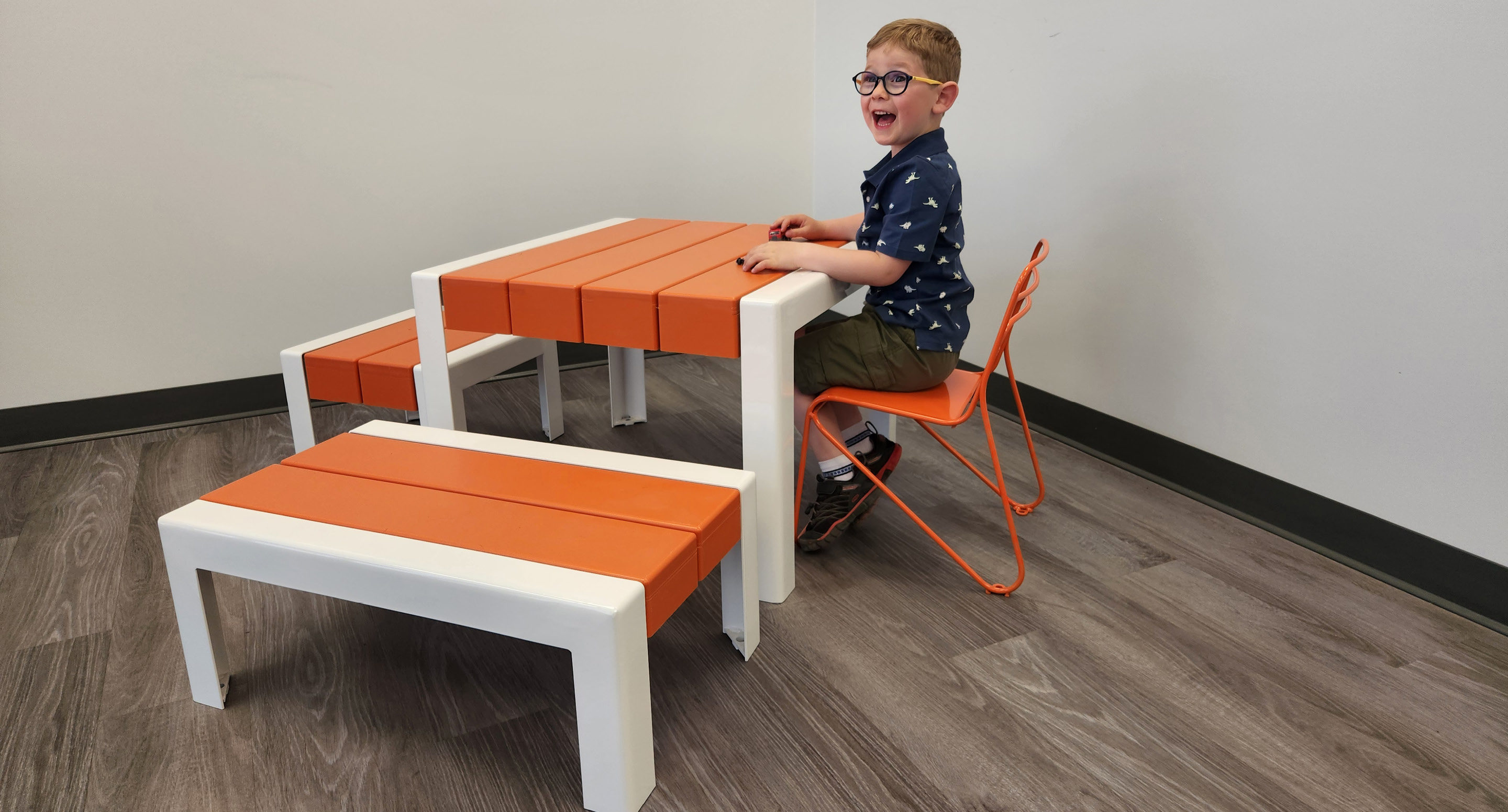 maglin_childrens-kontur-chair-3 Un jeune garçon sourit alors qu'il est assis à une table orange et blanche posée sur un sol en bois dans une pièce aux murs blancs.