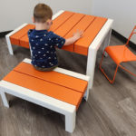 Un jeune enfant est assis sur un banc orange et blanc à une table assortie, avec une chaise métallique orange à proximité, dans une pièce au sol en bois.