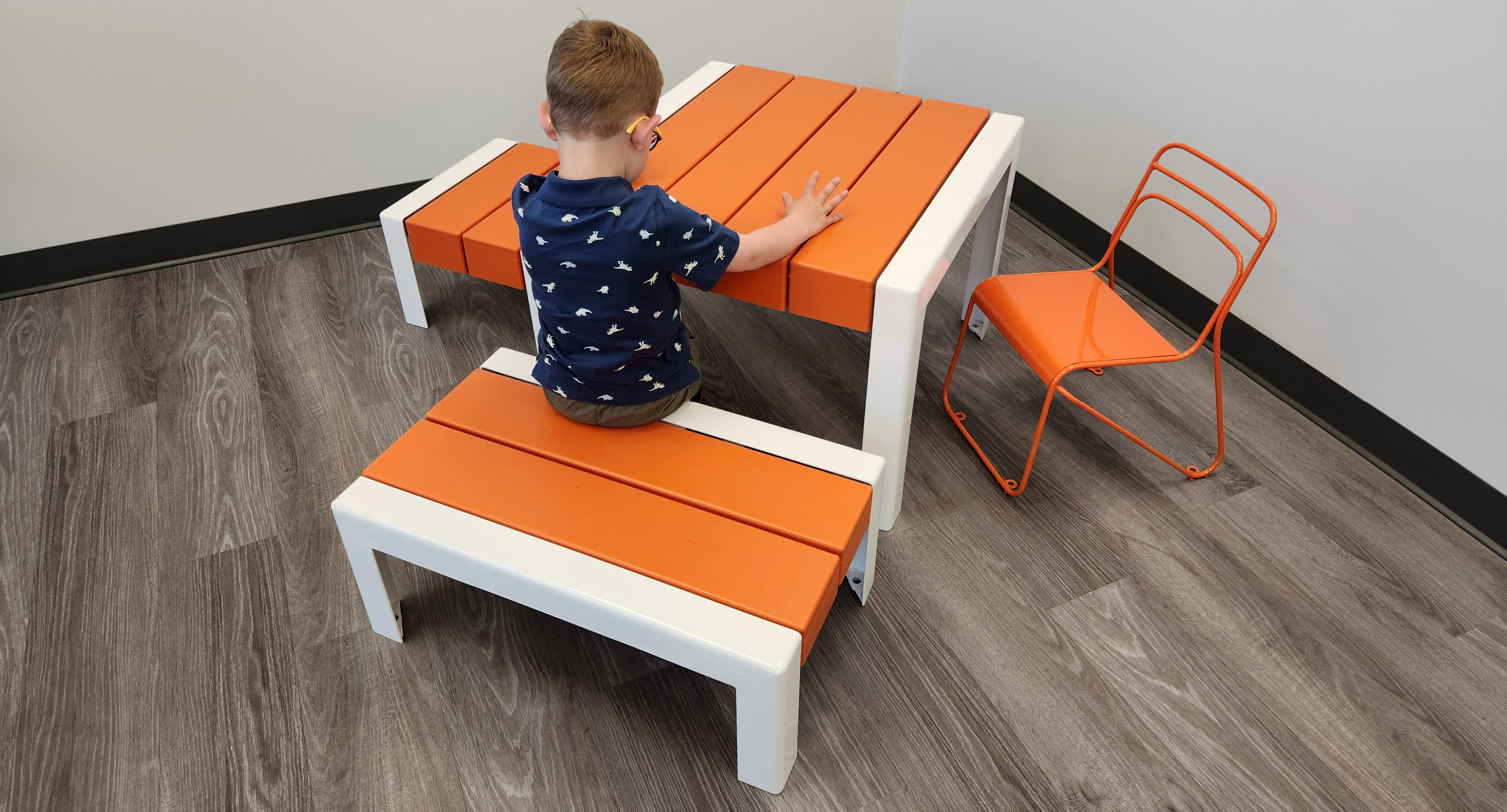 maglin_childrens-kontur-chair-4 Un jeune enfant est assis sur un banc orange et blanc à une table assortie, avec une chaise métallique orange à proximité, dans une pièce au sol en bois.