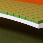 Vue rapprochée d'un banc moderne vert avec des pieds blancs sur une moquette verte et un fond orange.