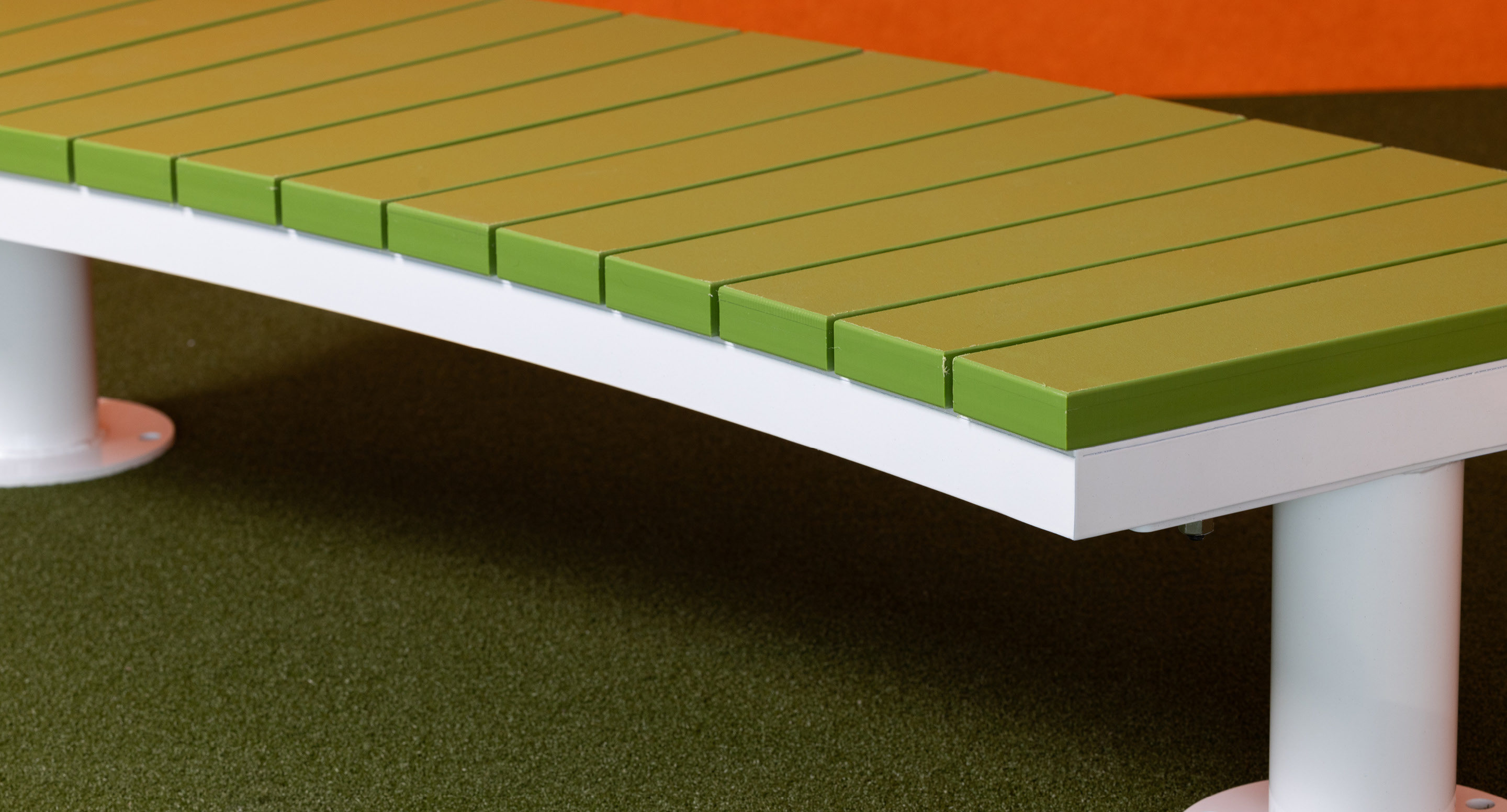 maglin_childrens-ogden-bench-1 Vue rapprochée d'un banc moderne vert avec des pieds blancs sur une moquette verte et un fond orange.