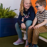 Deux enfants sont assis sur un banc vert, souriant et discutant, avec une grande fougère en pot et des murs orange et blancs en arrière-plan.