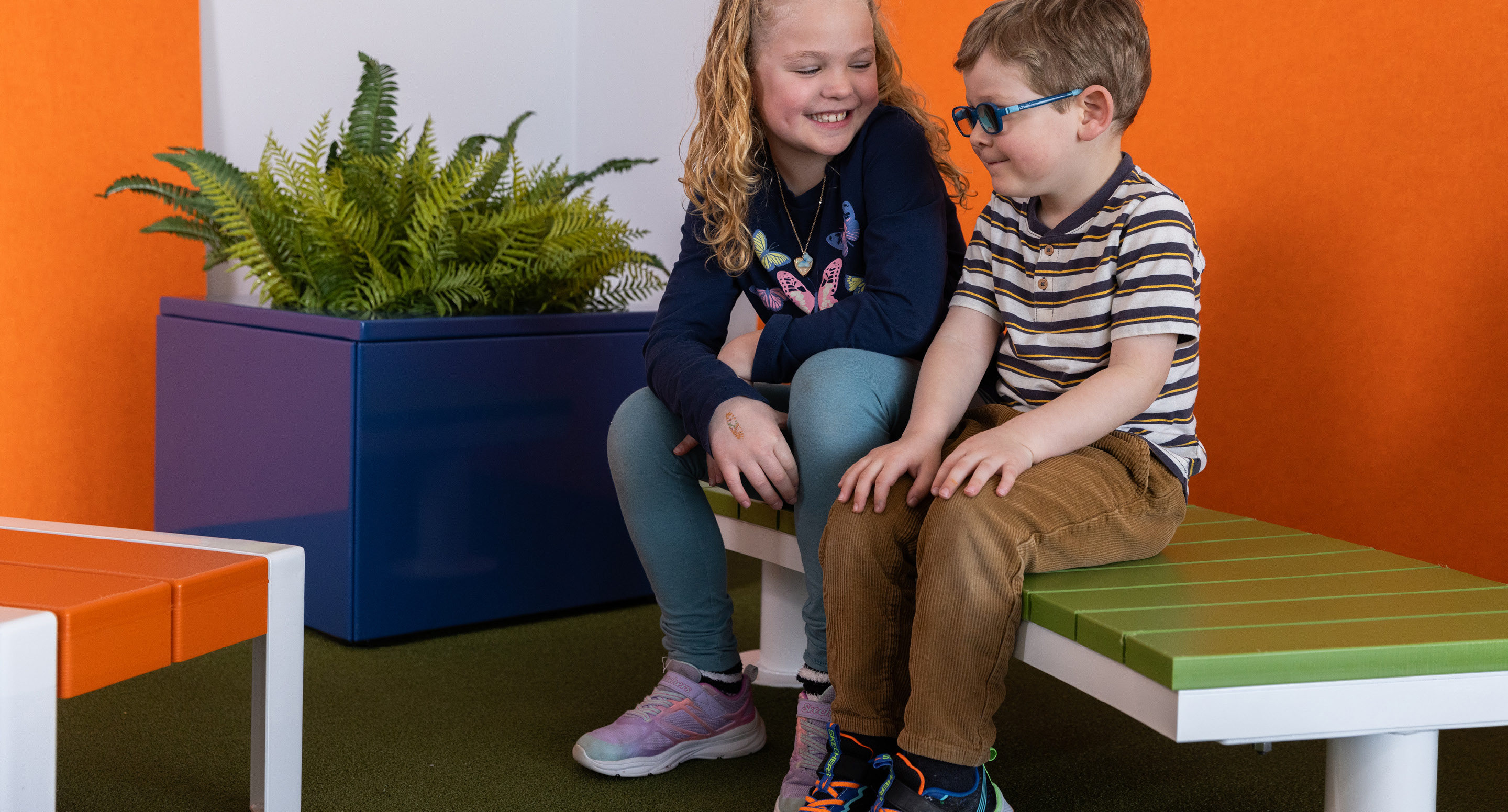 maglin_childrens-ogden-bench-2 Deux enfants sont assis sur un banc vert, souriant et discutant, avec une grande fougère en pot et des murs orange et blancs en arrière-plan.