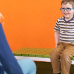 Un jeune garçon portant des lunettes et une chemise rayée est assis sur un banc vert, souriant, devant un mur orange. Un adulte est assis en face de lui, partiellement visible.