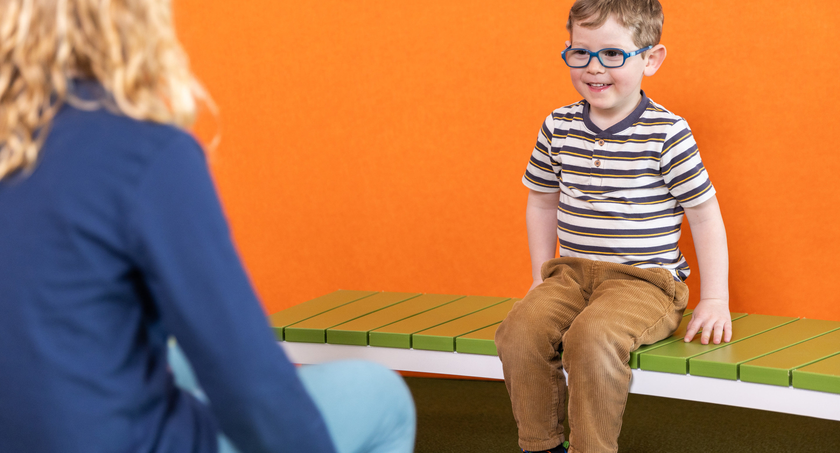maglin_childrens-ogden-bench-3 Un jeune garçon portant des lunettes et une chemise rayée est assis sur un banc vert, souriant, devant un mur orange. Un adulte est assis en face de lui, partiellement visible.