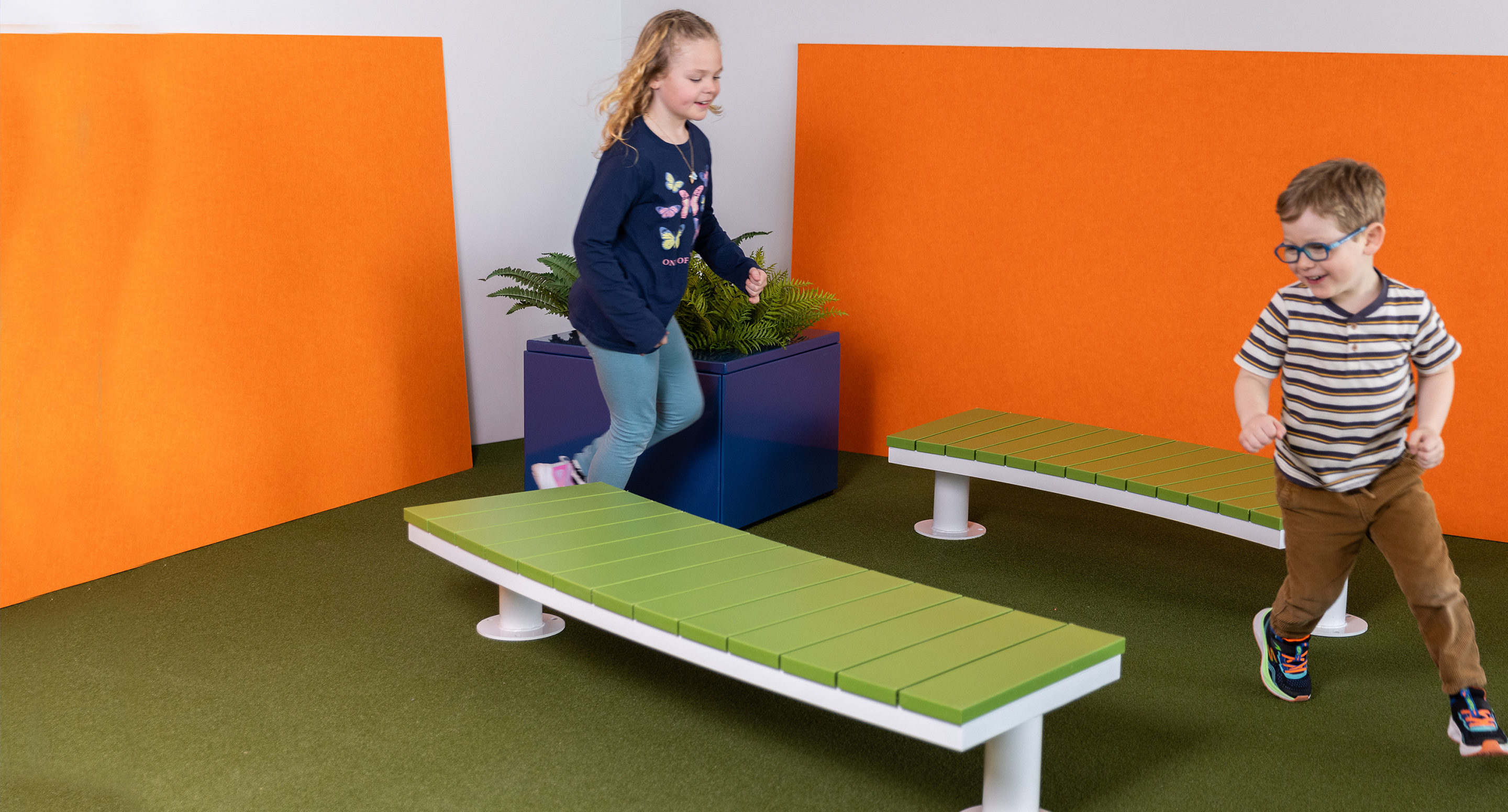 maglin_childrens-ogden-bench-4 Deux enfants jouent près de deux bancs verts sur du gazon artificiel, avec des panneaux orange et un pot de fleurs bleu en arrière-plan.