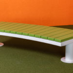 Un banc vert incurvé avec des pieds en métal est posé sur une moquette verte, contre des murs orange dans un coin.