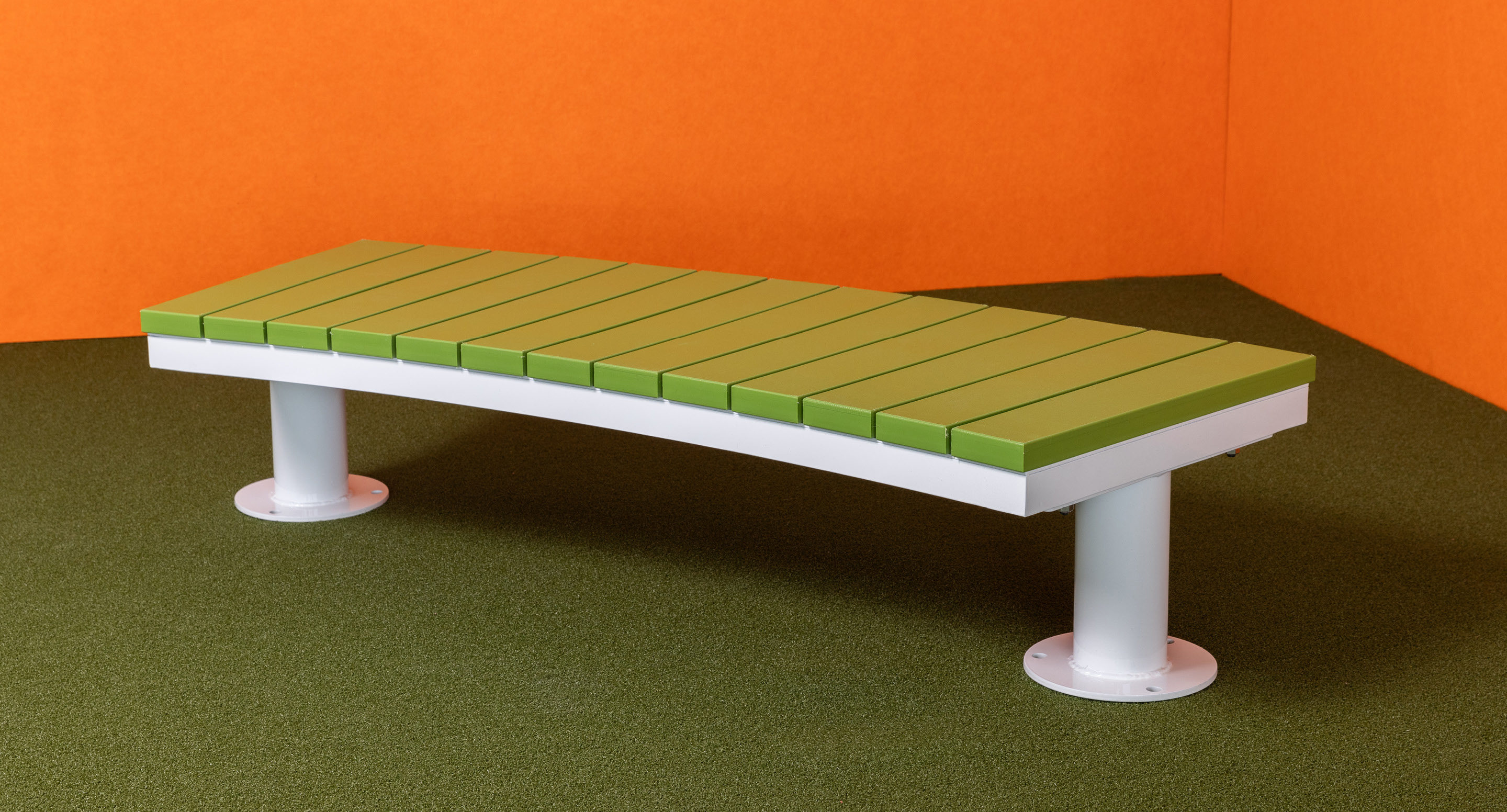 Un banc vert incurvé avec des pieds en métal est posé sur une moquette verte, contre des murs orange dans un coin.