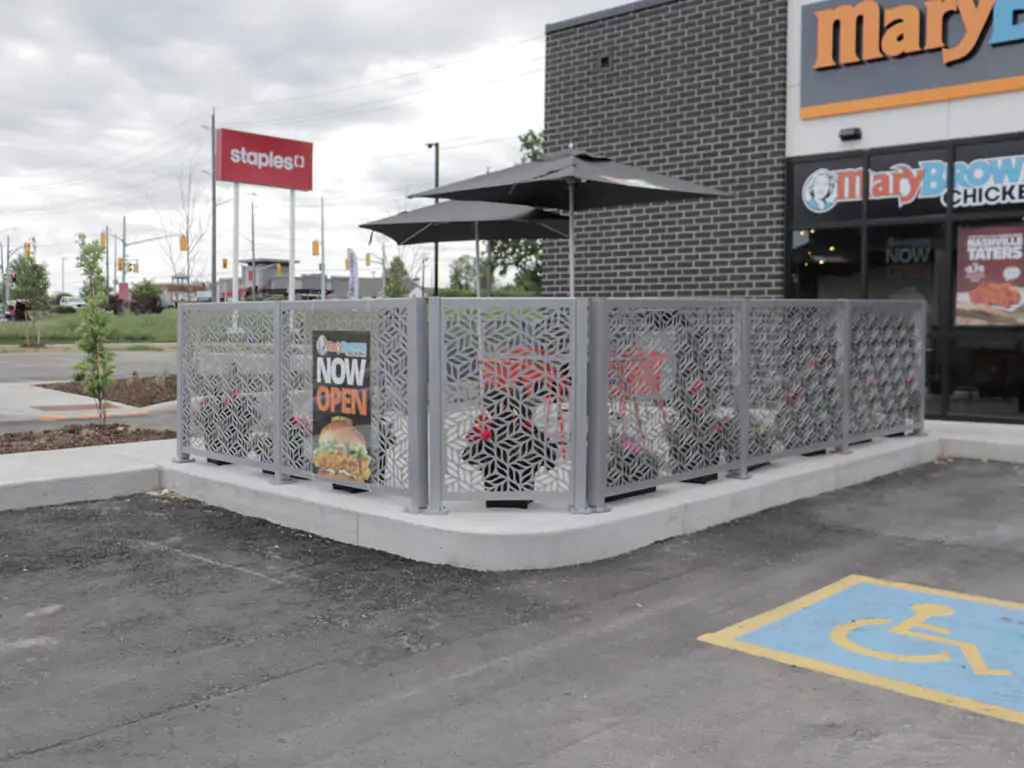 Outdoor restaurant patio with decorative metal fencing, potted plants, and an umbrella. A sign reads "Now Open" next to a handicap parking space in front of the building.
