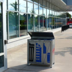Une poubelle extérieure divisée pour les déchets et le recyclage se trouve sur un trottoir à côté d'un bâtiment en verre, avec des voitures et un restaurant visibles à l'arrière-plan.