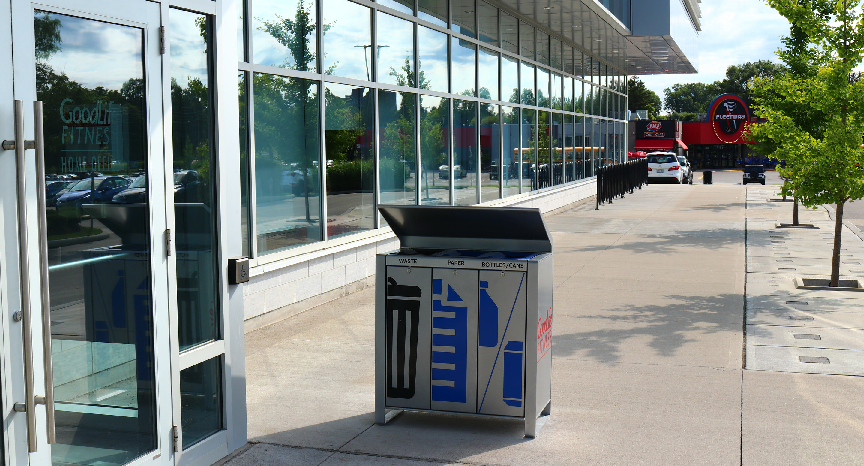 Une poubelle extérieure divisée pour les déchets et le recyclage se trouve sur un trottoir à côté d'un bâtiment en verre, avec des voitures et un restaurant visibles à l'arrière-plan.