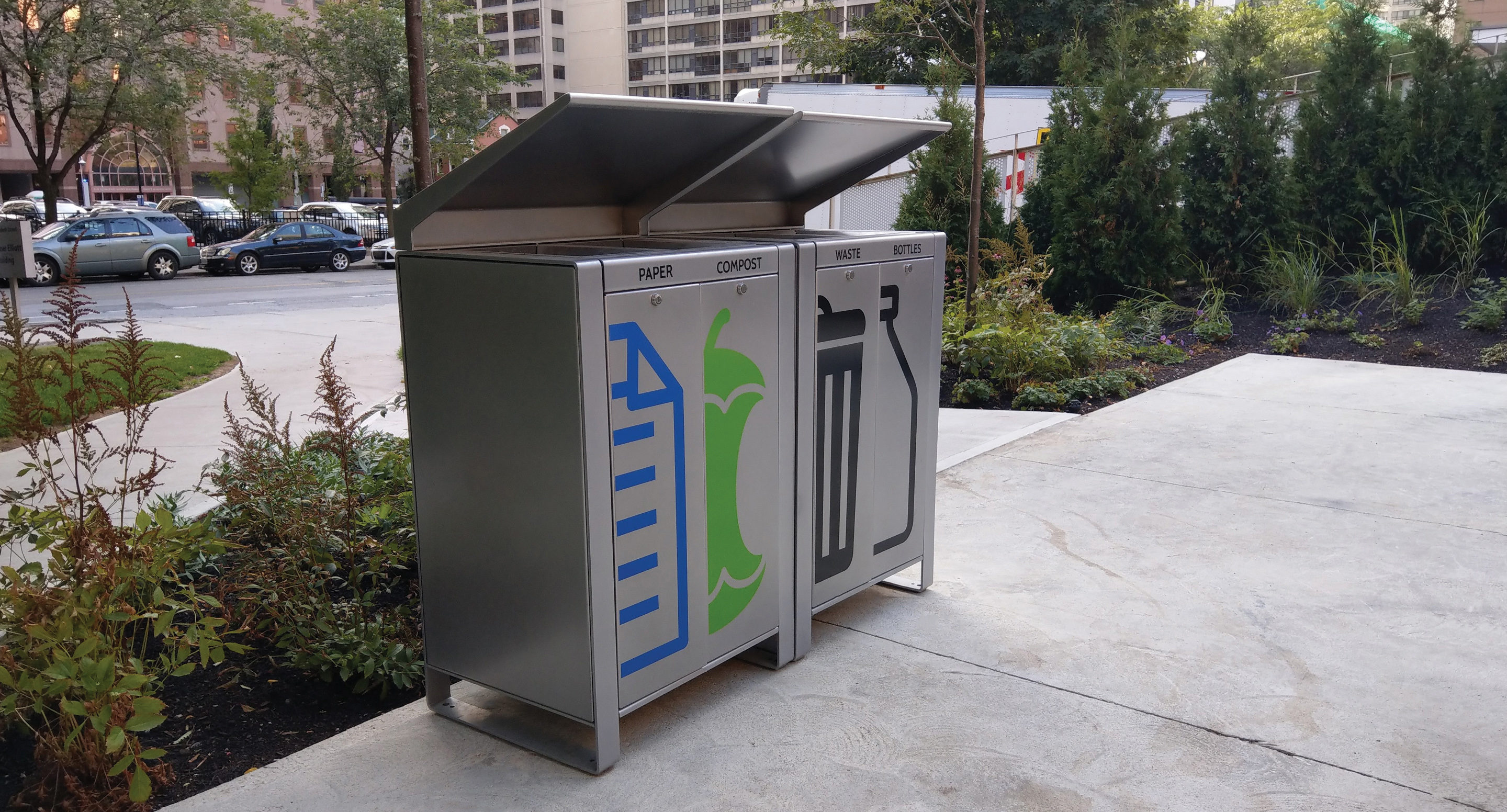 maglin_lexicon-recycle-receptacles-1 Trois poubelles extérieures étiquetées pour le papier, le compost et les déchets, placées sur un trottoir à proximité de verdure et de voitures garées dans une zone urbaine.