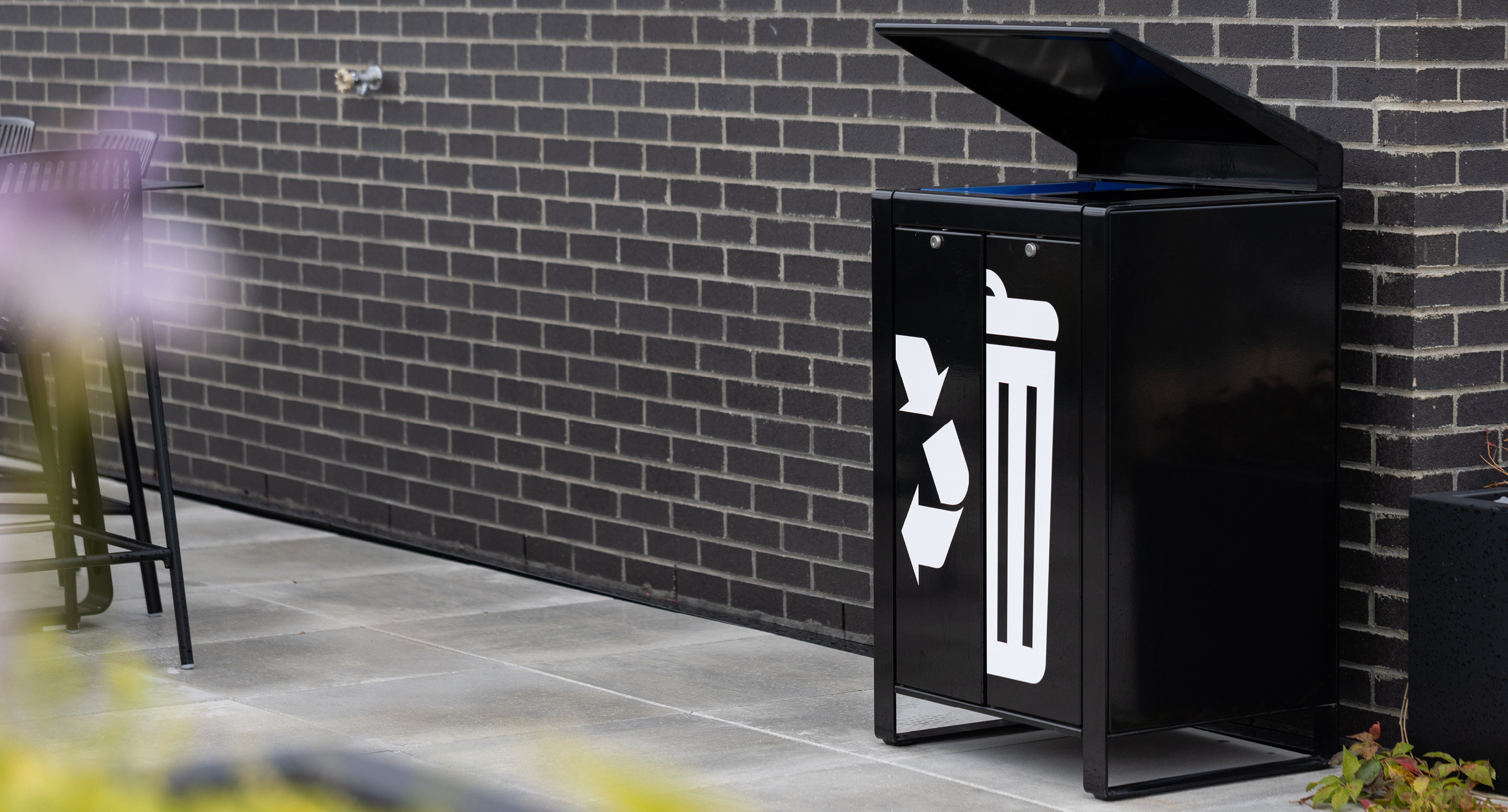 maglin_lexicon-recycle-receptacles-2 Une poubelle d'extérieur noire avec couvercle ouvert est posée contre un mur de briques sur un patio carrelé.