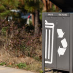 Poubelle extérieure pour les déchets et le recyclage avec des compartiments étiquetés et des icônes, située à côté d'un espace paysager avec de l'herbe et des buissons.