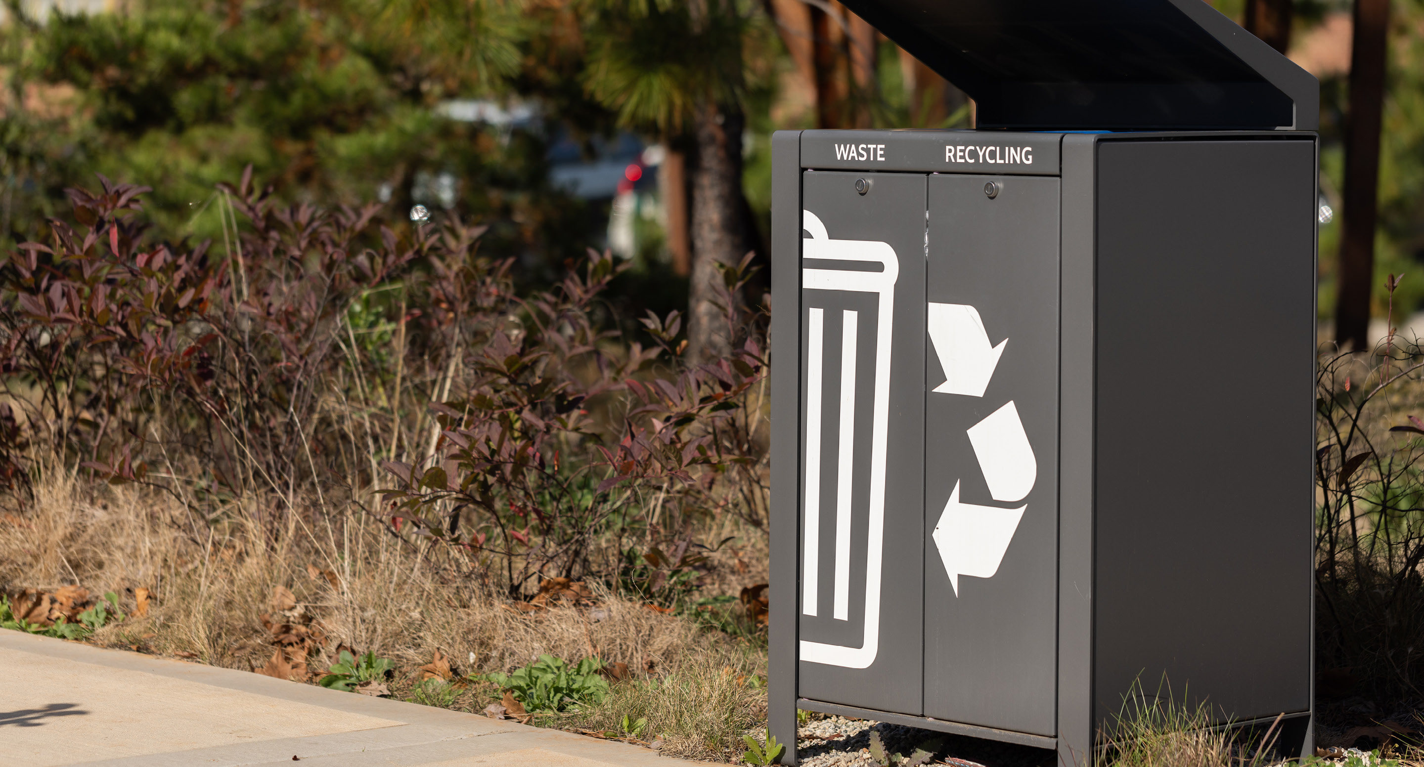 Poubelle extérieure pour les déchets et le recyclage avec des compartiments étiquetés et des icônes, située à côté d'un espace paysager avec de l'herbe et des buissons.