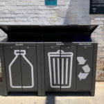 Trois poubelles noires avec de grandes icônes blanches pour le verre, les déchets et le recyclage sont placées contre un mur de briques à l'extérieur, sous la lumière du soleil.