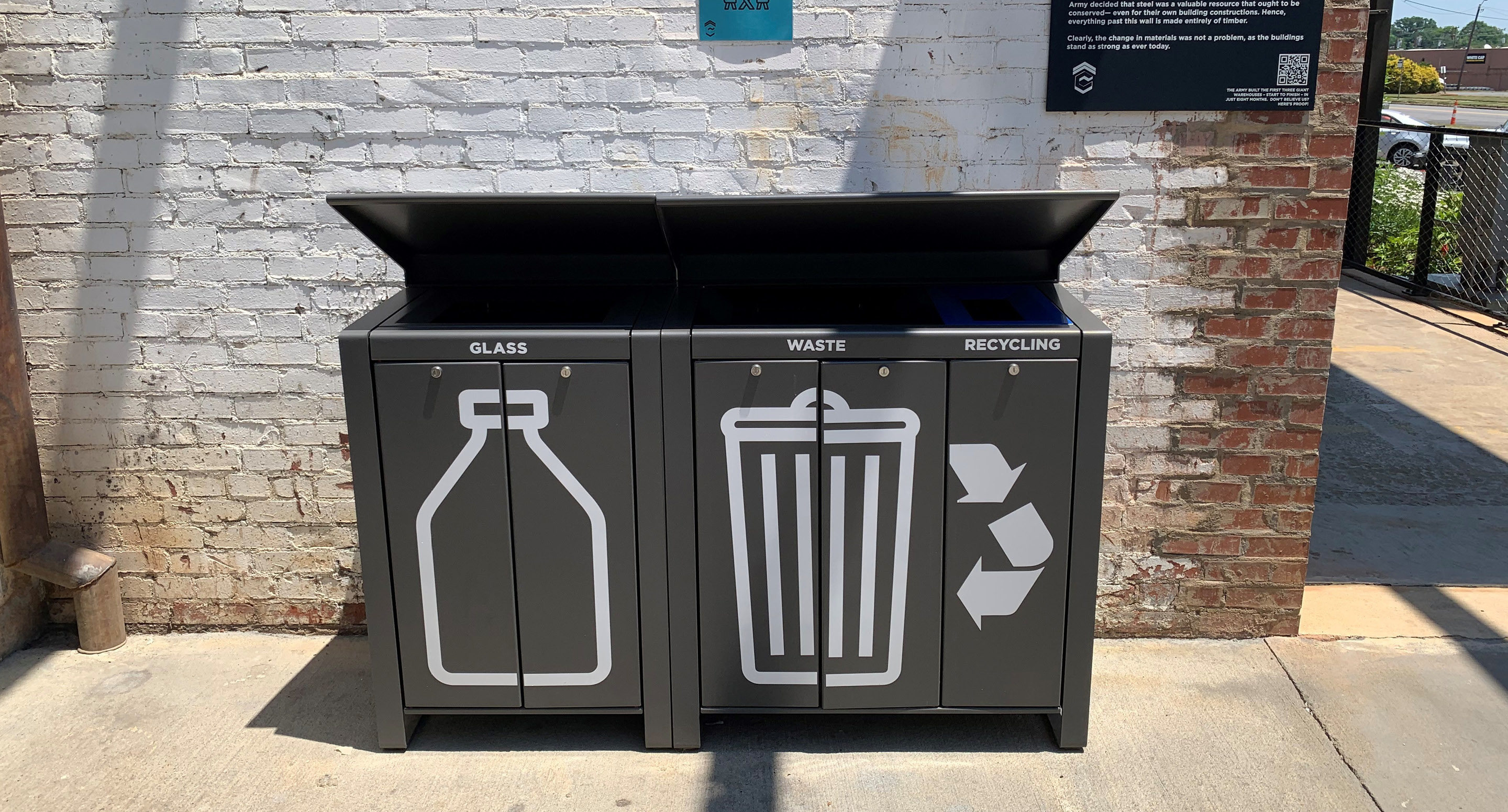 Trois poubelles noires avec de grandes icônes blanches pour le verre, les déchets et le recyclage sont placées contre un mur de briques à l'extérieur, sous la lumière du soleil.