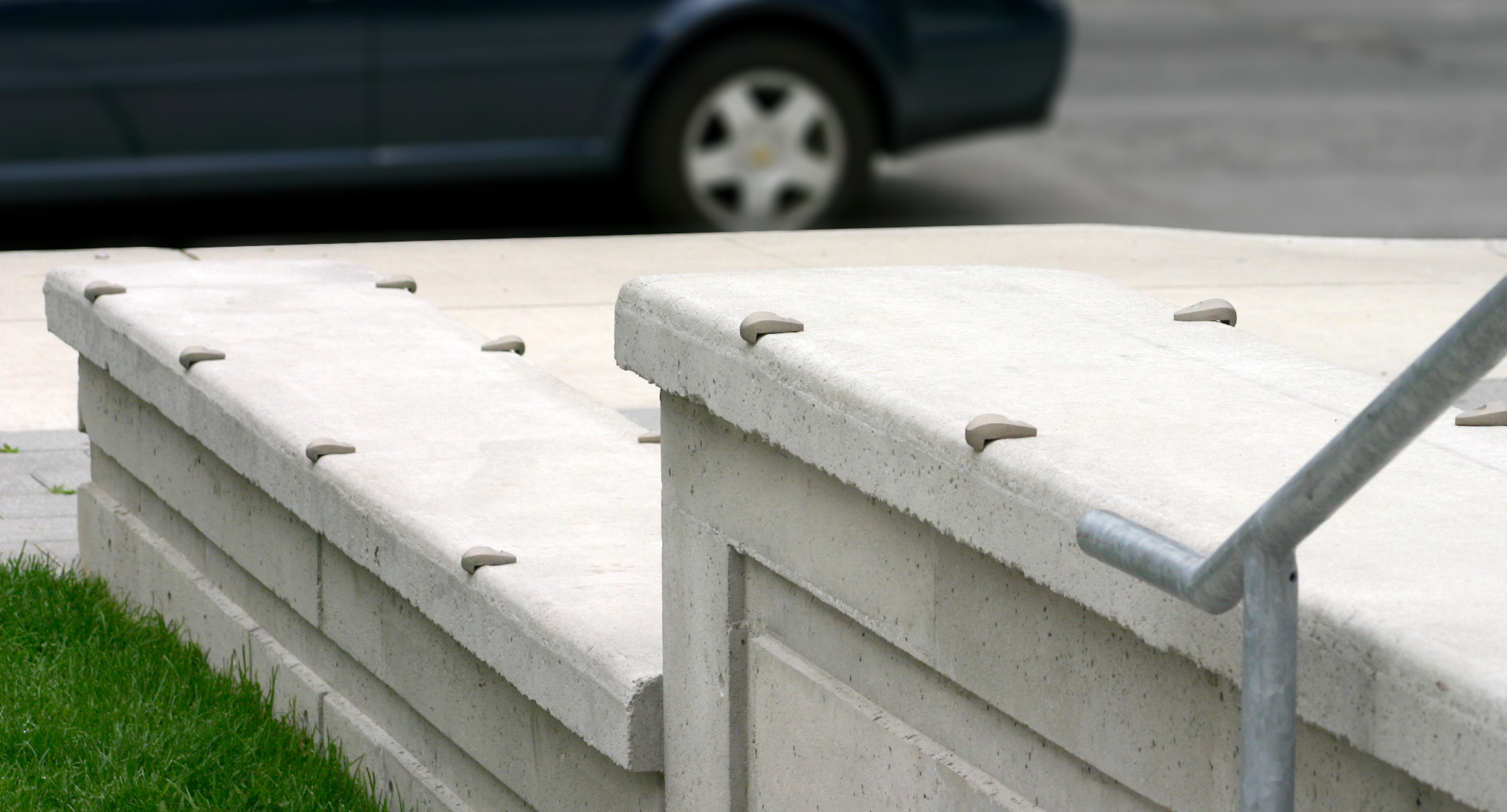 maglin_mac-0100-wall-mount-skate-deterrent Concrete ledges with metal skate-stoppers installed along the edges, next to a sidewalk and grass, with part of a parked car visible in the background.