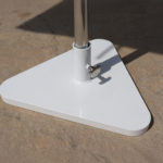 A close-up view of a white, triangular metal umbrella base with a vertical pole, set on a textured concrete surface.