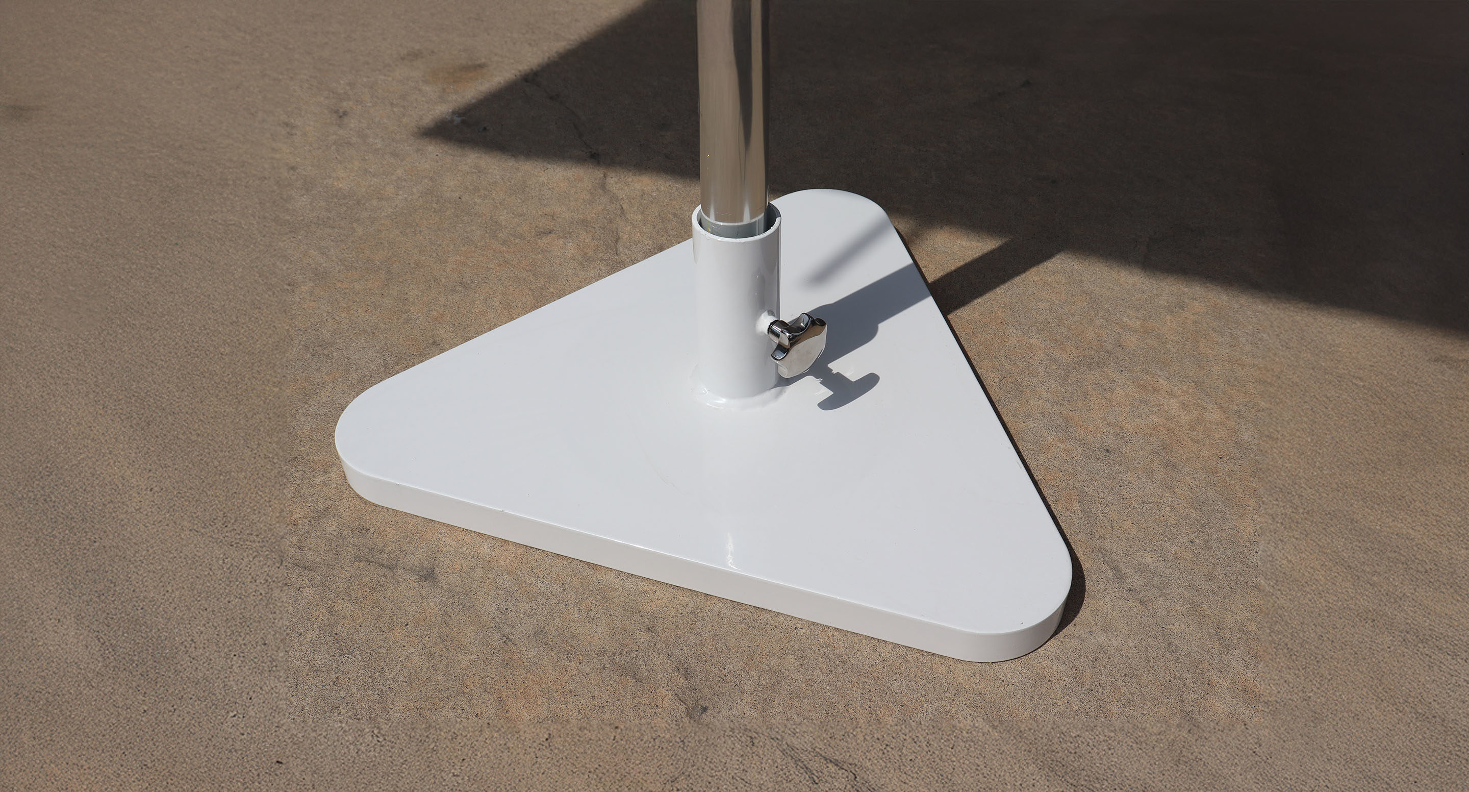 A close-up view of a white, triangular metal umbrella base with a vertical pole, set on a textured concrete surface.