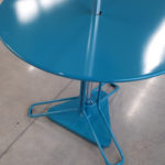 A round, blue metal table with a central pole and a triangular base stands on a polished concrete floor.