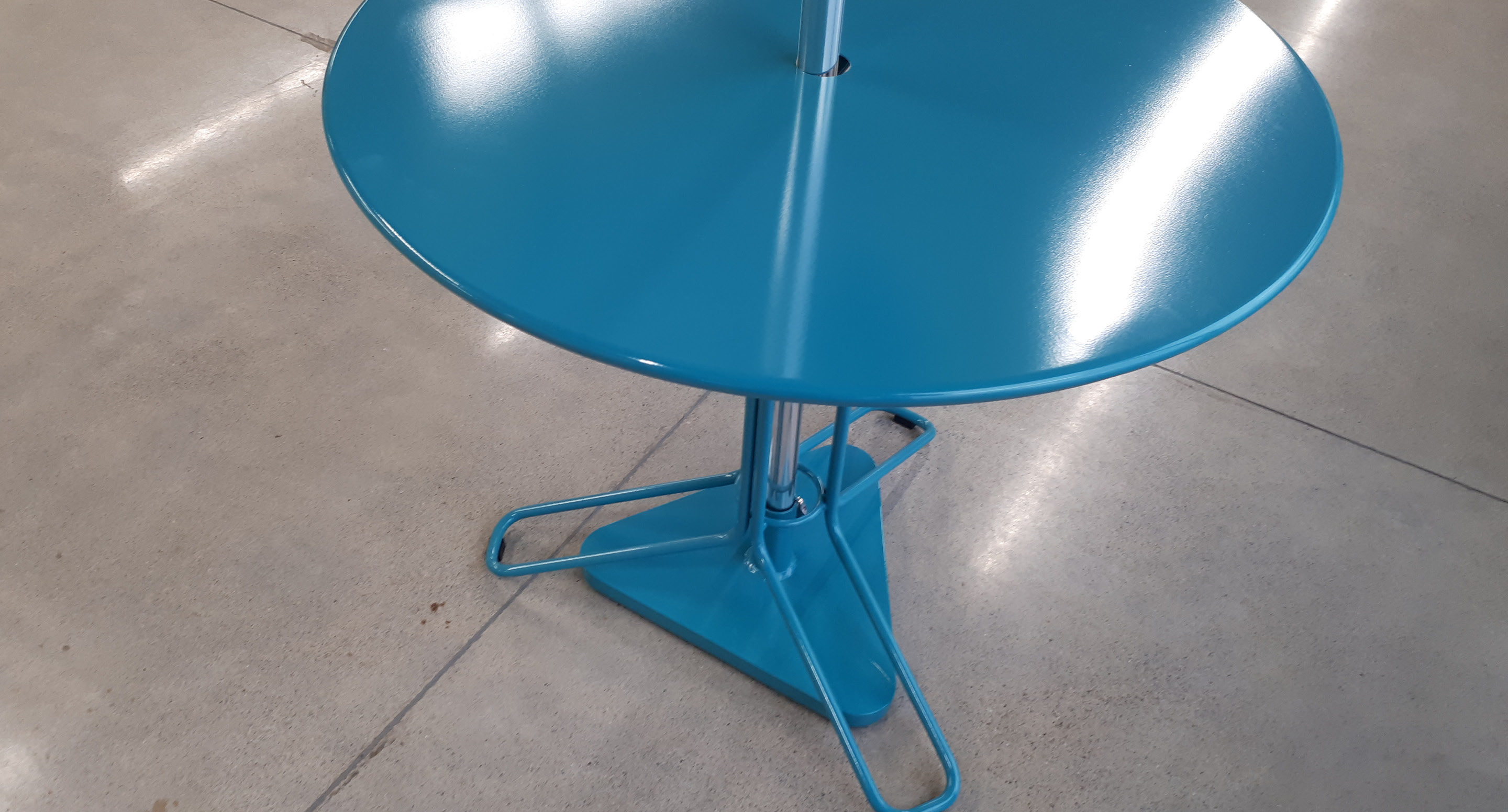 A round, blue metal table with a central pole and a triangular base stands on a polished concrete floor.