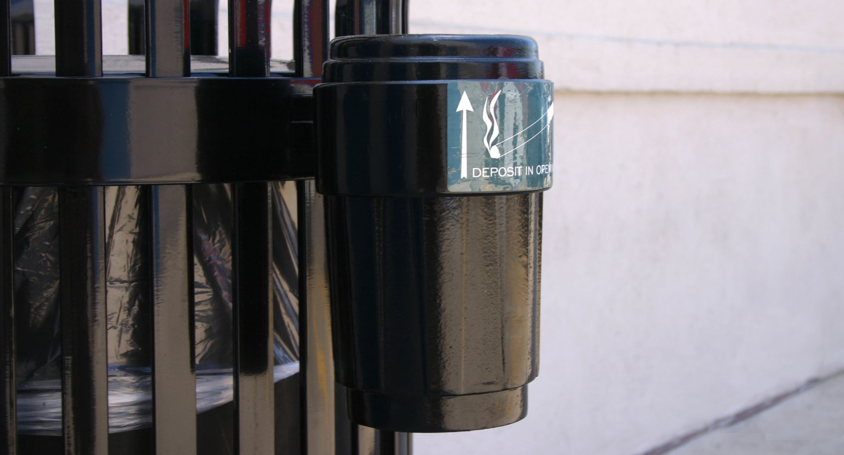 maglin_mar-0100-side-ash-receptacle-1 Une poubelle extérieure noire pour les cigarettes est fixée à une rambarde métallique, avec une étiquette indiquant où déposer les déchets de cigarettes.