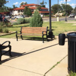 Deux bancs en bois et deux poubelles sont placés le long d'un trottoir dans un parc aménagé avec de l'herbe, des arbres et des lampadaires.