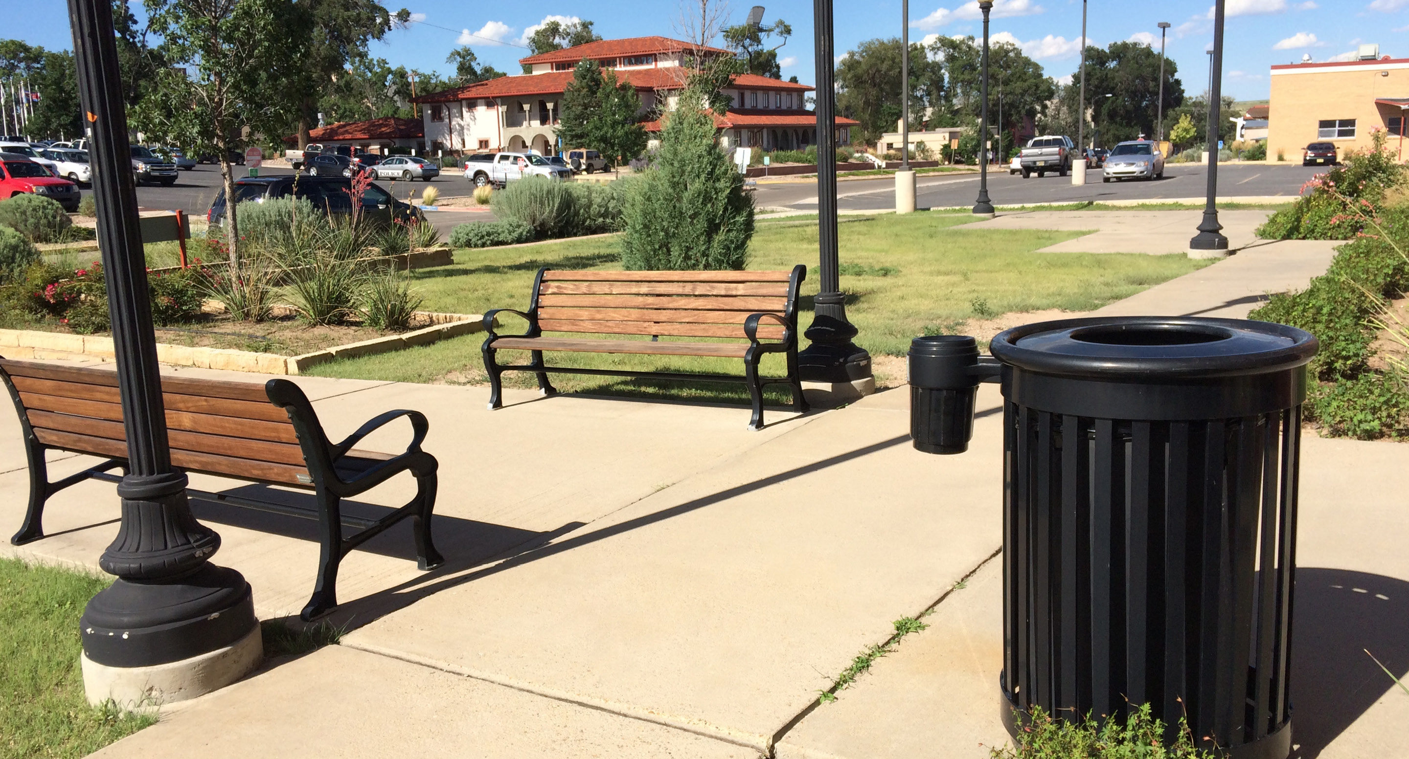 Deux bancs en bois et deux poubelles sont placés le long d'un trottoir dans un parc aménagé avec de l'herbe, des arbres et des lampadaires.