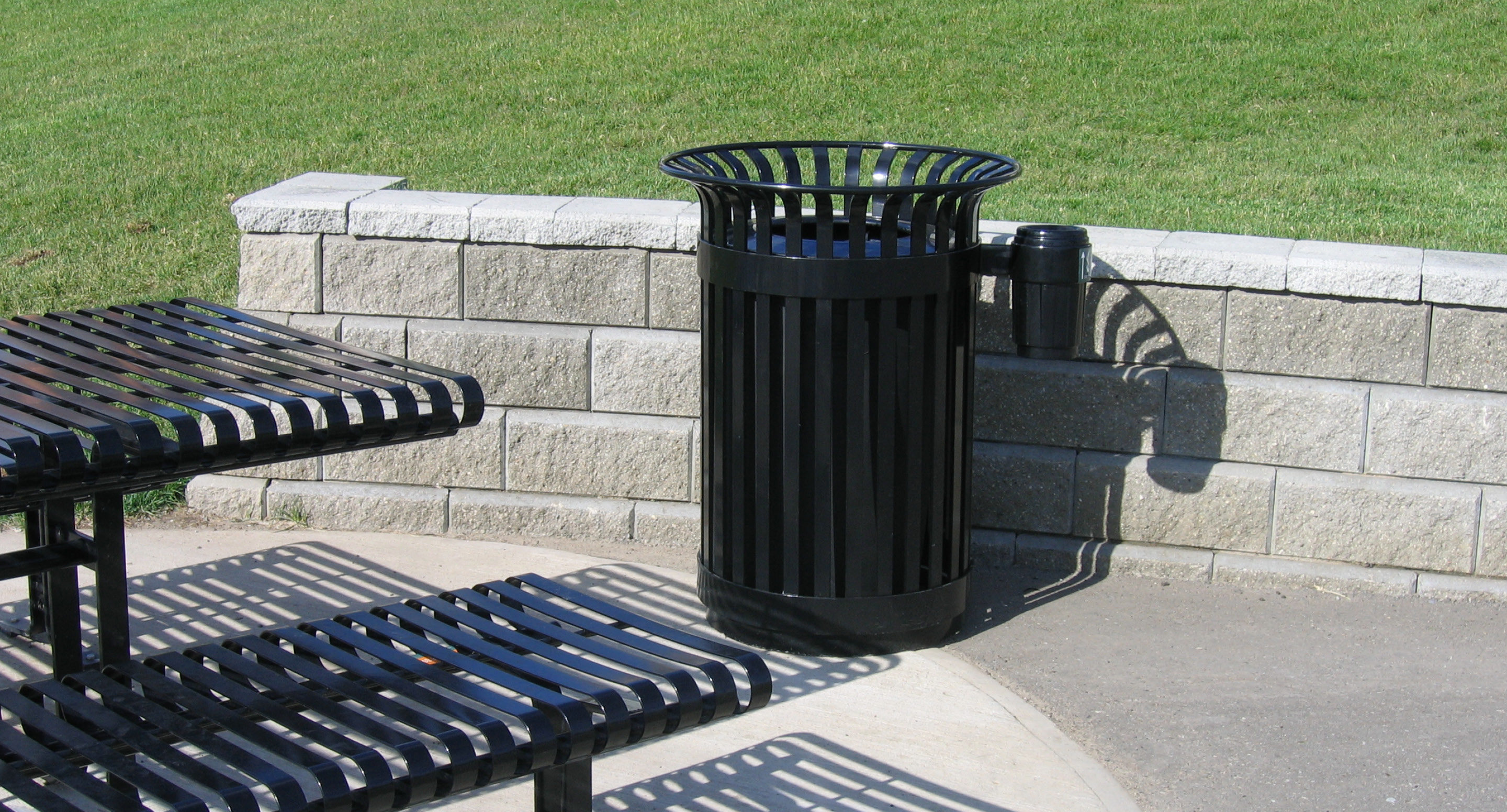 maglin_mar-0100-side-ash-receptacle-3 Poubelle en métal noir à côté d'un banc assorti et d'un petit mur en pierre dans un parc avec de l'herbe verte et un chemin pavé.
