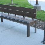 A brown metal bench on a concrete sidewalk next to a cigarette disposal post and a grassy area.