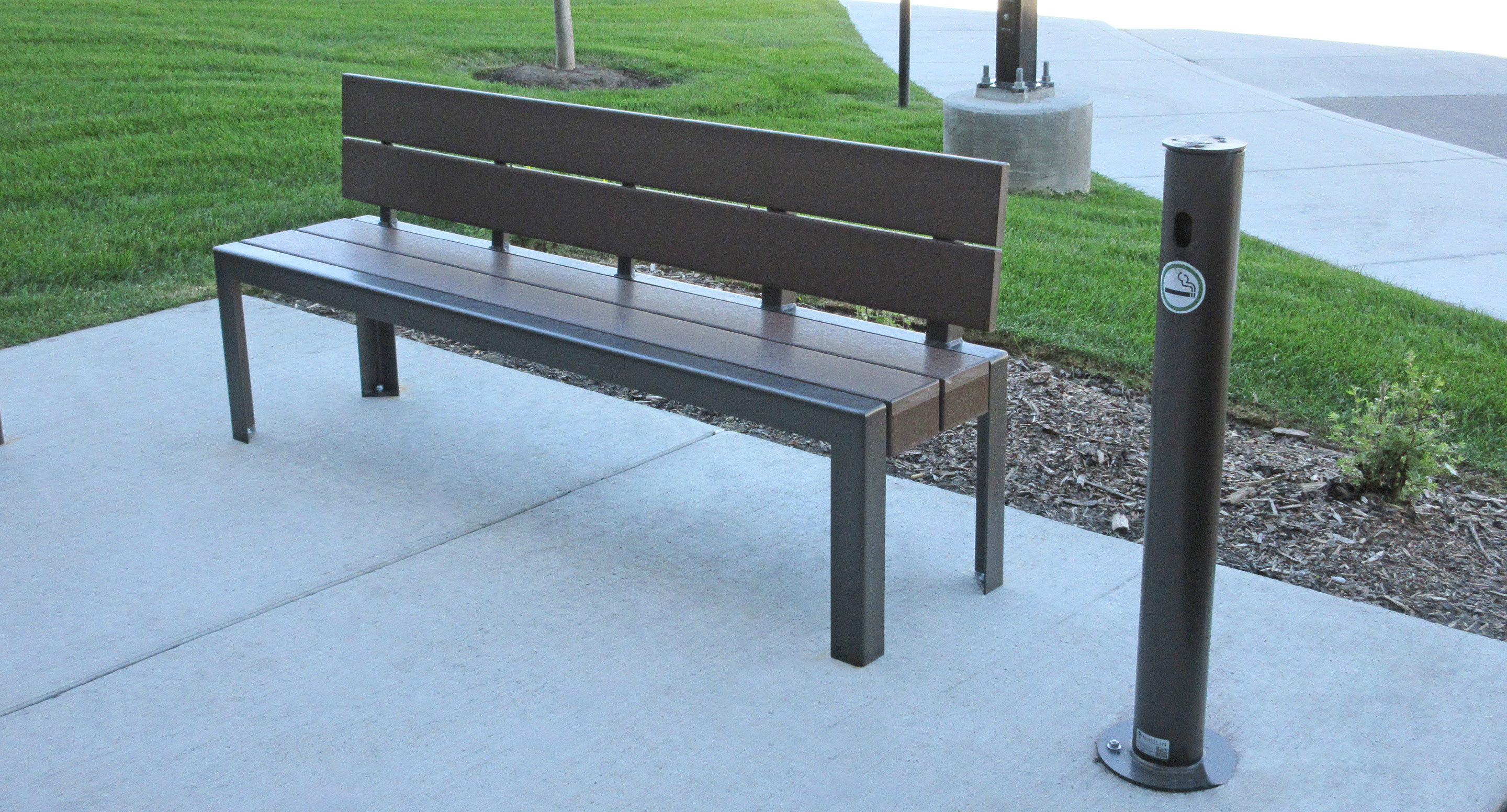 maglin_mar-0300-ash-receptacle-1 A brown metal bench on a concrete sidewalk next to a cigarette disposal post and a grassy area.