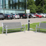 Two empty gray benches face each other beside a black trash can near a parking lot with several parked cars and a modern glass building in the background.