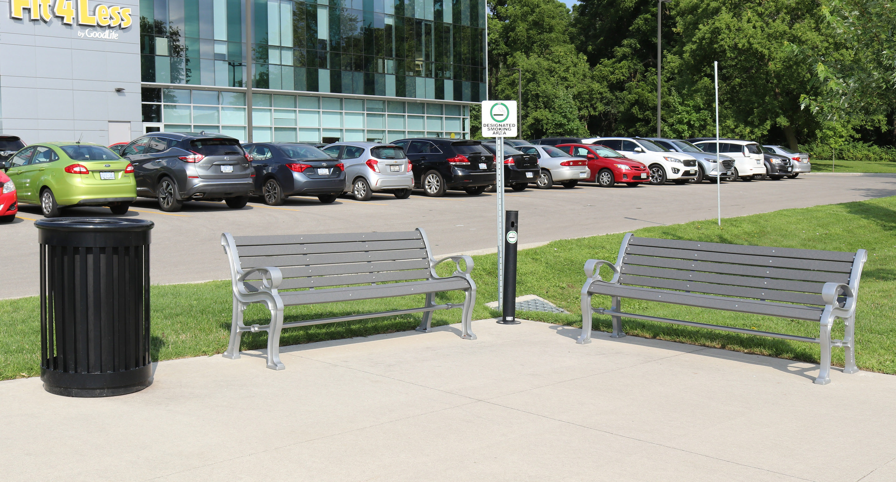 Two empty gray benches face each other beside a black trash can near a parking lot with several parked cars and a modern glass building in the background.