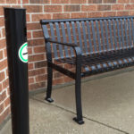 A black metal bench is placed on a concrete sidewalk against a red brick wall; a black post with a green and white sticker stands nearby.