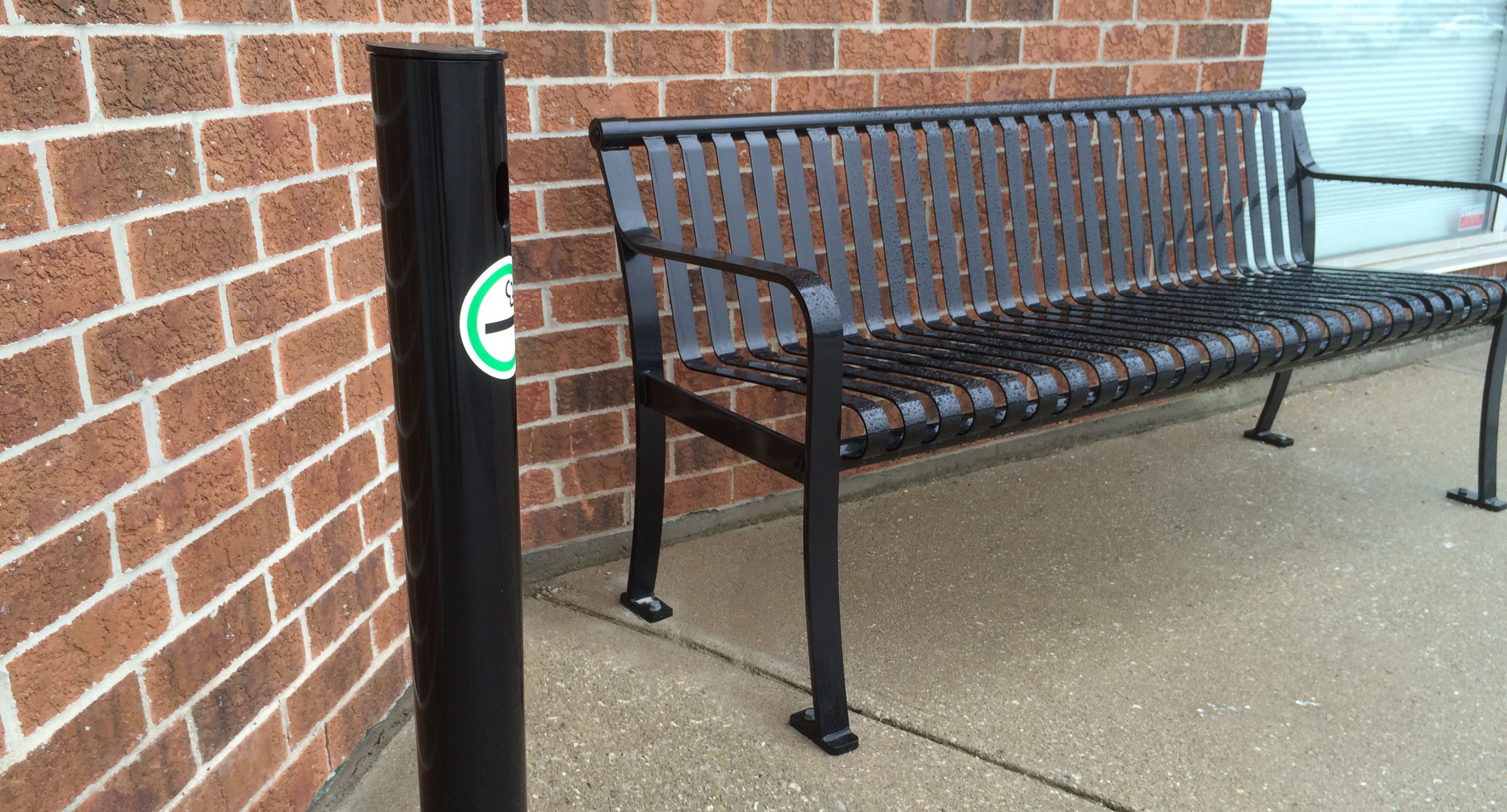 maglin_mar-0300-ash-receptacle-3 A black metal bench is placed on a concrete sidewalk against a red brick wall; a black post with a green and white sticker stands nearby.