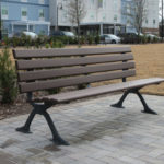 A brown wooden park bench with metal legs sits on a paved area near a grassy field, with shrubs and buildings in the background.